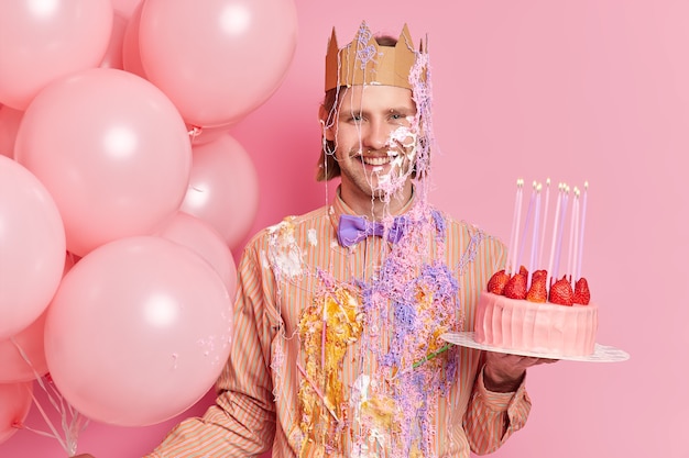 Knappe vrolijke man staat vuil met room houdt heerlijke aardbeientaart viert verjaardag stands met vakantieattributen geïsoleerd over roze muur