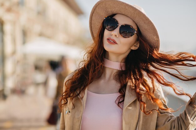 Knappe vrouw in zwarte zonnebril speelt met rood krullend haar