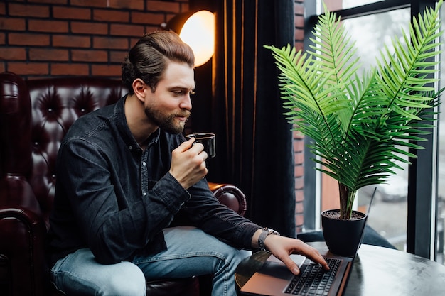 Knappe zakenman die op laptop werkt, kopje koffie of latte vasthoudt in modern café.