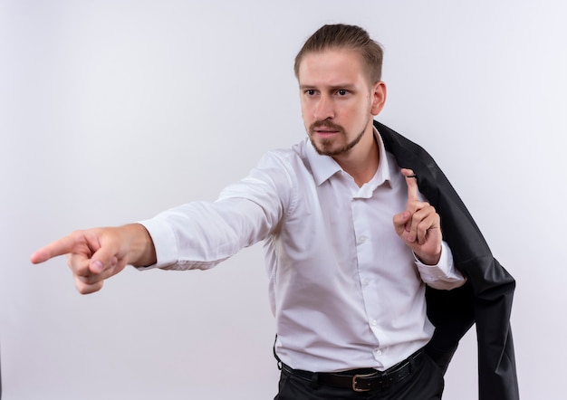 Knappe zakenman die zijn jasje op schouder draagt die met vinger aan de kant met ernstig gezicht richt dat zich over witte achtergrond bevindt