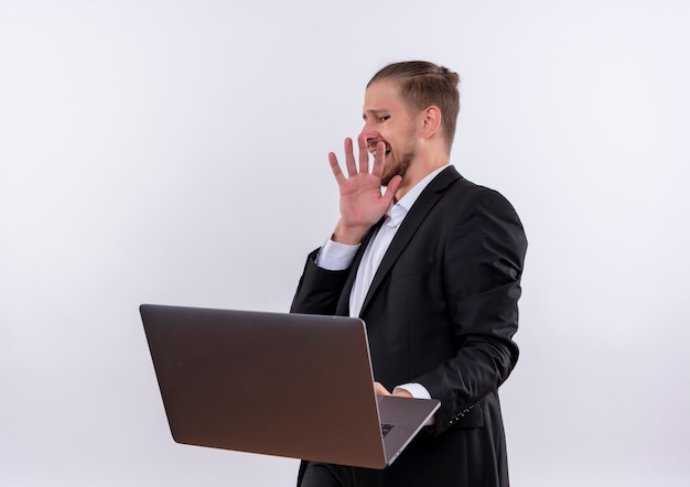 Knappe zakenman dragen pak met laptop computer maken defensie gebaar met walgen expressie staande op witte achtergrond
