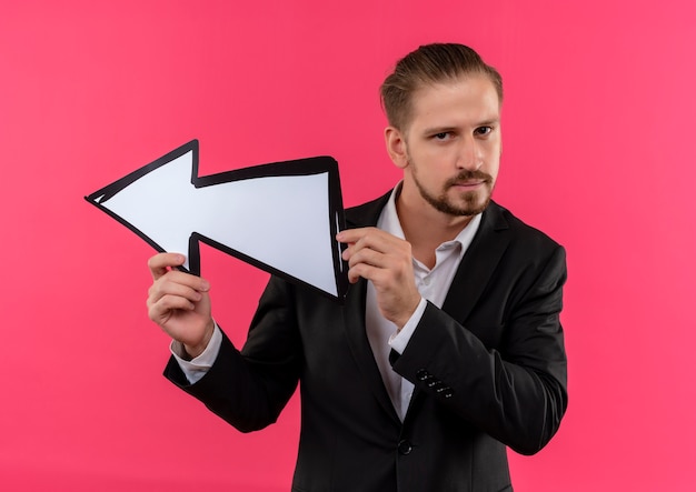 Gratis foto knappe zakenman dragen pak met pijl teken wijst met het naar links kijken camera met ernstig gezicht staande over roze achtergrond