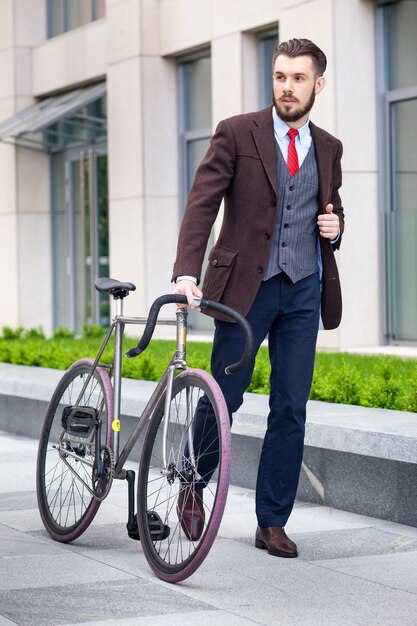 Knappe zakenman in een jas en een rode stropdas en zijn fiets op stadsstraten. Het concept van de moderne levensstijl van jonge mannen