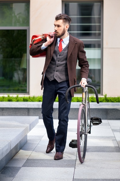 Knappe zakenman in een jas en een rode stropdas en zijn fiets op stadsstraten. Het concept van de moderne levensstijl van jonge mannen
