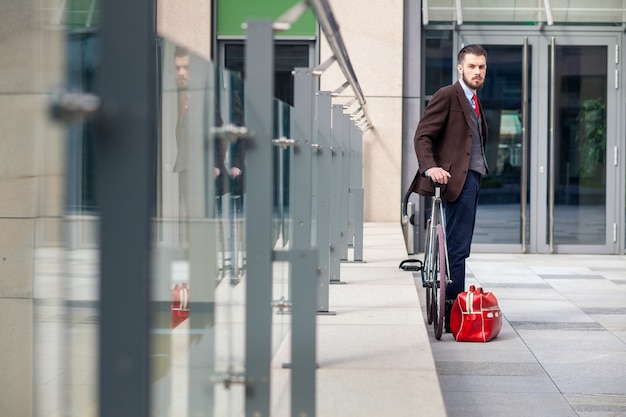 Knappe zakenman in een jas en een rode stropdas en zijn fiets op stadsstraten. rode zak ligt naast. Het concept van de moderne levensstijl van jonge mannen