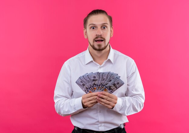 Knappe zakenman in wit overhemd die contant geld tonen dat verbaasd status over roze achtergrond kijkt