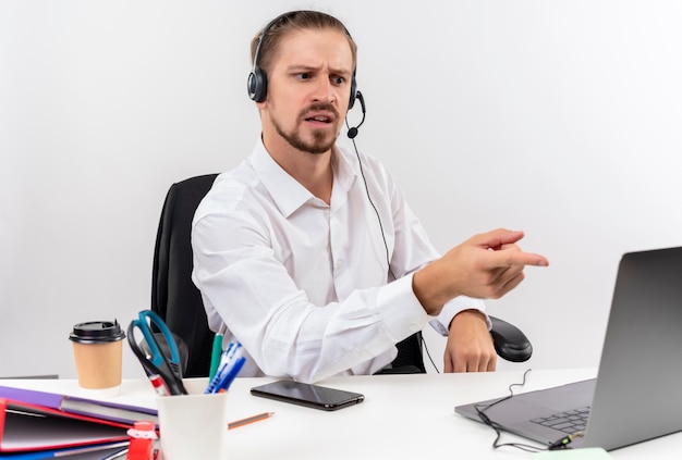 Knappe zakenman in wit overhemd en koptelefoon met een microfoon die op laptop werkt op zoek verward zittend aan tafel in offise op witte achtergrond