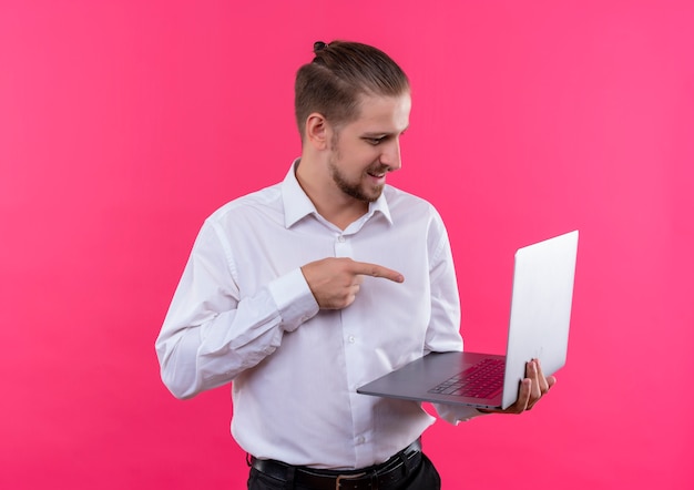 Knappe zakenman in wit overhemd met laptop wijzend met vinger naar laptop scherm verward staande over roze achtergrond