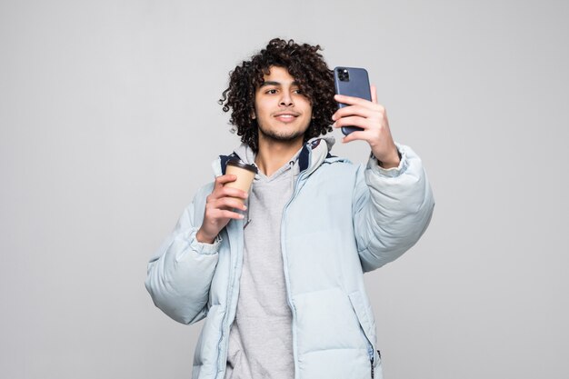 Knappe zekere jonge krullende mens die een selfie nemen, die kop van meeneemkoffie status houden geïsoleerd over grijze muur houden