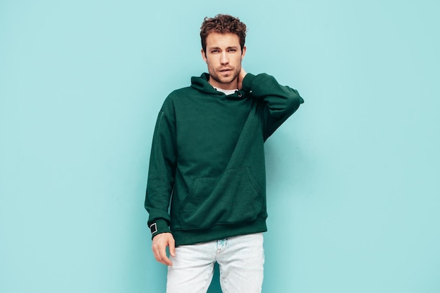 Knappe zelfverzekerde hipster modelSexy ongeschoren man gekleed in zomer stijlvolle groene hoodie en jeans kleding Mode man met krullend kapsel poseren in studio geïsoleerd op blauw