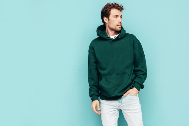 Knappe zelfverzekerde hipster modelSexy ongeschoren man gekleed in zomer stijlvolle groene hoodie en jeans kleding Mode man met krullend kapsel poseren in studio geïsoleerd op blauw