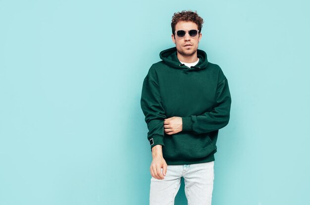 Knappe zelfverzekerde hipster modelSexy ongeschoren man gekleed in zomer stijlvolle groene hoodie en jeans kleding Mode man met krullend kapsel poseren in studio geïsoleerd op blauw