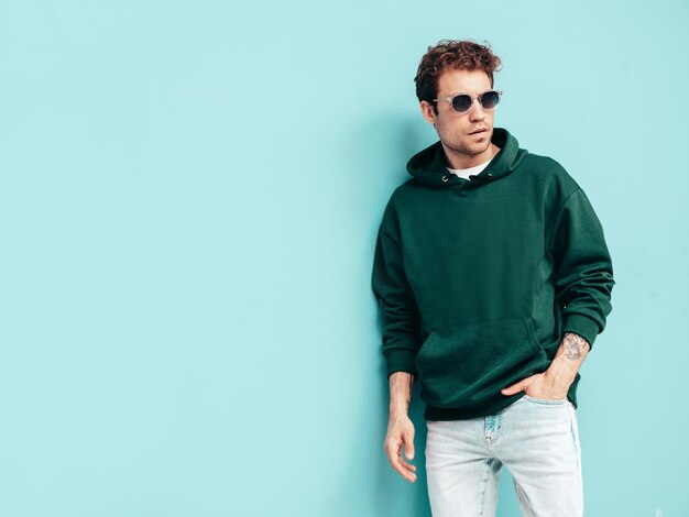Knappe zelfverzekerde hipster modelSexy ongeschoren man gekleed in zomer stijlvolle groene hoodie en jeans kleding Mode man met krullend kapsel poseren in studio geïsoleerd op blauw