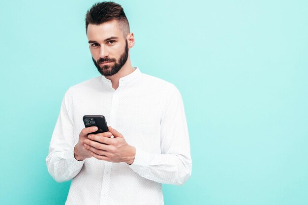 Knappe zelfverzekerde modelSexy stijlvolle man gekleed in shirt en spijkerbroek Mode hipster man poseren in de buurt van blauwe muur in studio Holding smartphone kijken naar mobiele telefoon scherm met behulp van apps