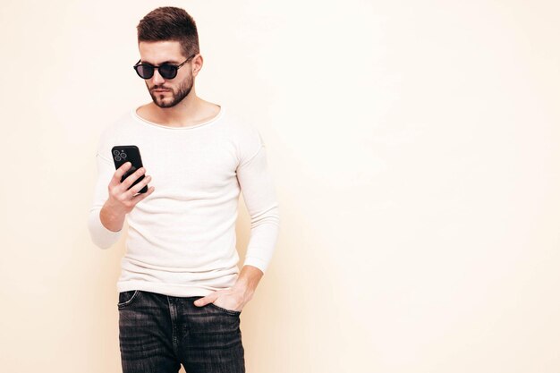 Knappe zelfverzekerde modelSexy stijlvolle man gekleed in trui en spijkerbroek Mode hipster man poseren in de buurt van witte muur in studio Holding smartphone kijken naar mobiel scherm met behulp van apps geïsoleerd