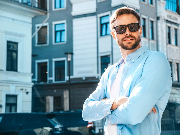 Knappe zelfverzekerde stijlvolle hipster lamberseksueel model moderne man gekleed in blauw shirt mode man poseren in de buurt van wolkenkrabber op de straat achtergrond in zonnebril buiten bij zonsondergang gekruiste armen