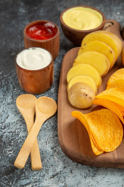 Knapperige chips en ongekookte aardappelen op houten snijplank en verschillende kruiden op grijze tafel