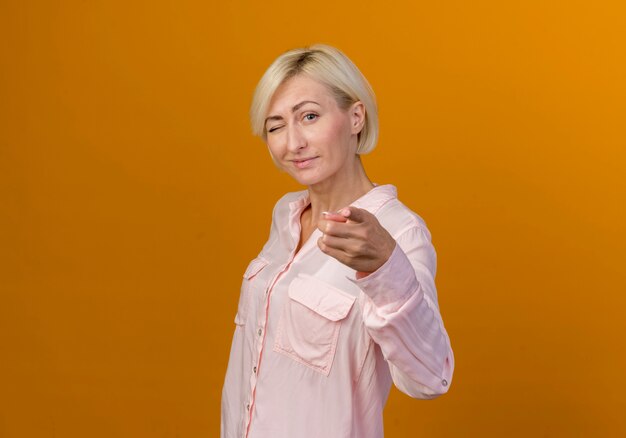 Knipperde jonge blonde Slavische vrouw die je gebaar toont dat op oranje muur wordt geïsoleerd