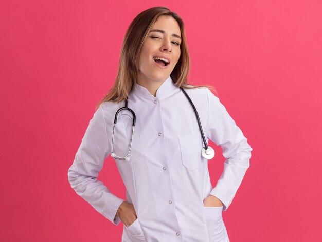 Knipperde jonge vrouwelijke arts die medische mantel met een stethoscoop aanbrengt dient de zak in die op roze muur wordt geïsoleerd
