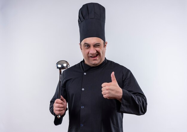 Knipperde mannelijke kok van middelbare leeftijd in eenvormige chef-kok met spatel zijn duim omhoog op geïsoleerde witte muur