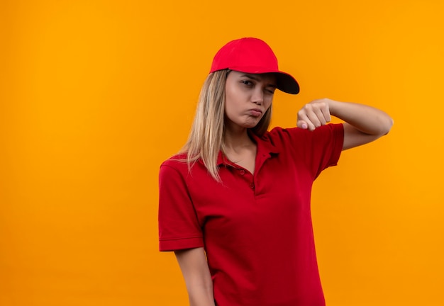 Knipperend jong leveringsmeisje die rood uniform en GLB dragen die vuist opheffen die op oranje achtergrond wordt geïsoleerd