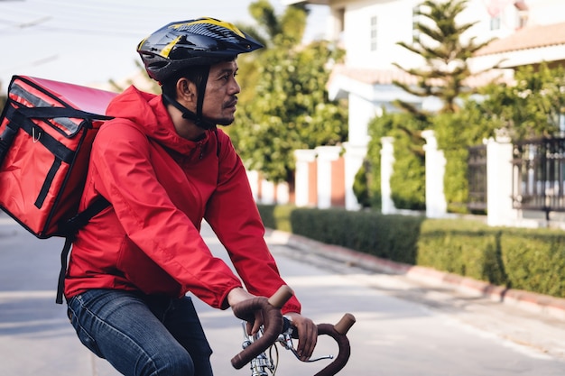 Koerier in rood uniform met een bezorgdoos op de fiets en op de mobiele telefoon kijkend om het adres te controleren om eten bij de klant te bezorgen. koerier op een fiets die voedsel in de stad bezorgt.