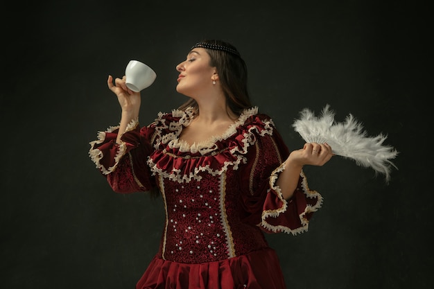 Koffie drinken, houdt een donzige ventilator vast. middeleeuwse jonge vrouw in rode vintage kleding op donkere achtergrond. vrouwelijk model als hertogin, koninklijk persoon. concept vergelijking van tijdperken, modern, mode, schoonheid.