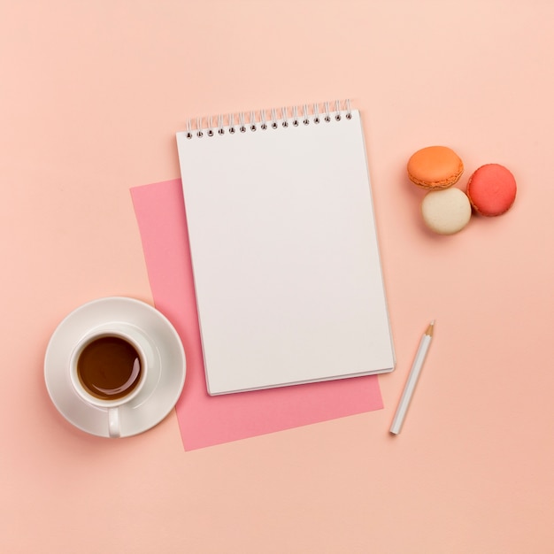 Koffiekop met spiraalvormige blocnote, wit potlood en makarons op gekleurde achtergrond
