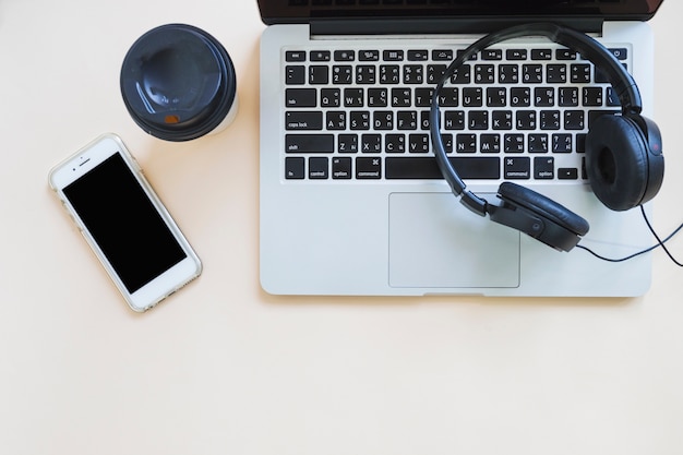 Koffiekop; mobiel en hoofdtelefoon over de laptop tegen beige achtergrond