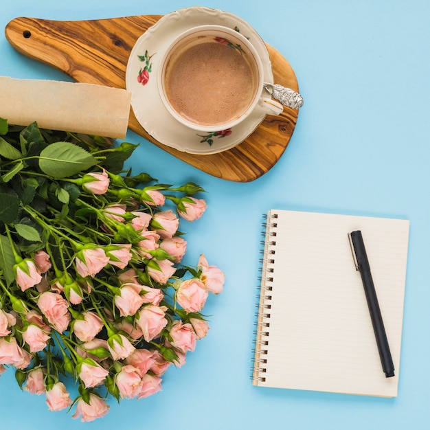 Koffiekop; roze rozen; spiraal notitieblok; pen op blauwe achtergrond