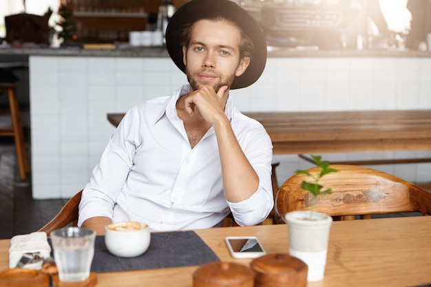 Koffietijd. modieuze jonge hipster in zwarte hoed rust in café, zittend aan tafel met kopje cappuccino en generieke mobiele telefoon, hand op zijn kin, kijken en glimlachen