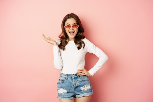Koket en stijlvol brunette meisje, lachen en glimlachen, het dragen van een zonnebril, het dragen van witte blouse en jeans, roze achtergrond. Ruimte kopiëren