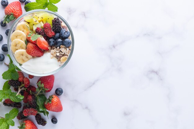 Kom havergranola met yoghurt, verse bosbessen, moerbei, aardbeien, kiwi, banaan, munt en notenbord voor gezond ontbijt, bovenaanzicht, kopieerruimte, plat leggen. vegetarisch voedselconcept.