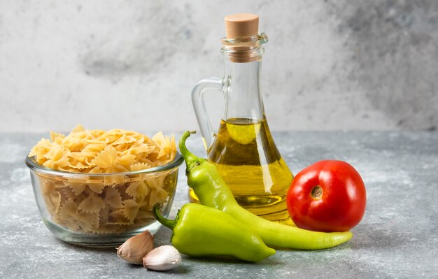 Kom rauwe farfalledeegwaren met fles olijfolie en groenten op marmeren lijst.