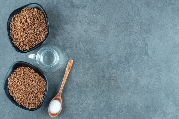 Kookopstelling van twee kommen boekweit, een kleine kan water en een lepel zout op marmeren achtergrond. hoge kwaliteit foto