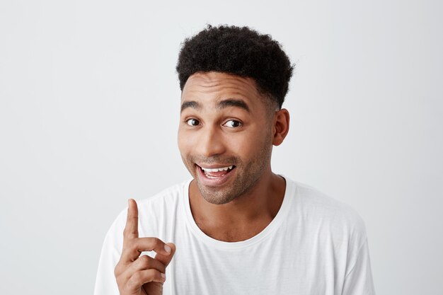 Kopieer ruimte. Portret van jonge aantrekkelijke zwarte huid vrolijke man met afro kapsel in casual t-shirt lachend met tanden, wijzend ondersteboven met vinger, op zoek in de camera met gelukkige uitdrukking