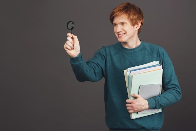 Kopieer ruimte. Portret van jonge mooie vrolijke mannelijke student lachend met tanden, met veel papieren en vergrootglas voor hem, opzij op zoek met opgewonden en gelukkige uitdrukking