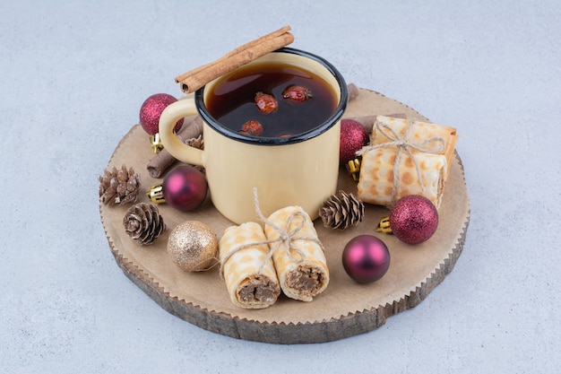 Kopje thee met rozenbottels, koekjes en kerstballen op een houten bord.