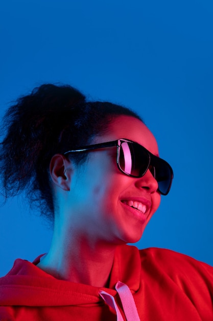 Koptelefoon. Portret van de Afro-Amerikaanse vrouw geïsoleerd op blauwe studio achtergrond in veelkleurige neonlicht. Mooi vrouwelijk model. Concept van menselijke emoties, gezichtsuitdrukking, verkoop, advertentie, mode.