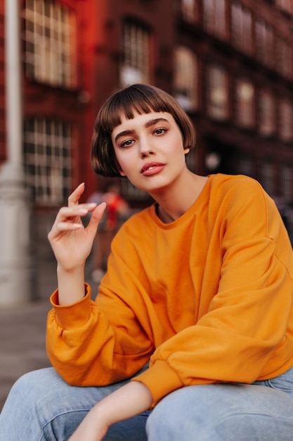 Kortharig meisje in oranje sweatshirt poseert buiten Mooie vrouw in stijlvolle lichte outfit kijkt in de camera