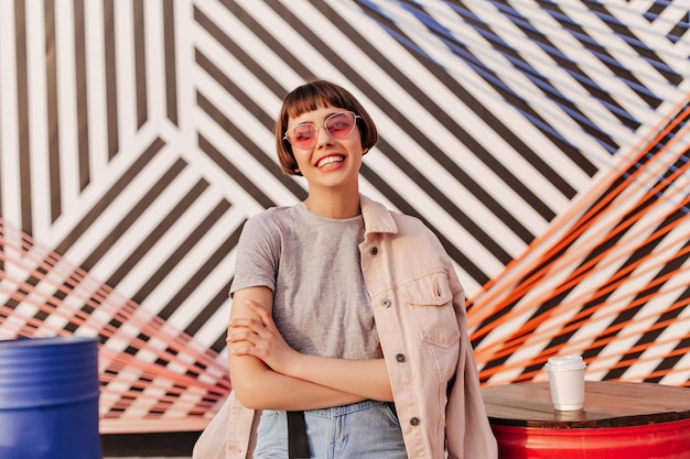 Kortharig meisje met roze zonnebril glimlachend in gestreepte achtergrond Tiener met donkerbruin haar in grijze t-shirt en beige jas poseren in café