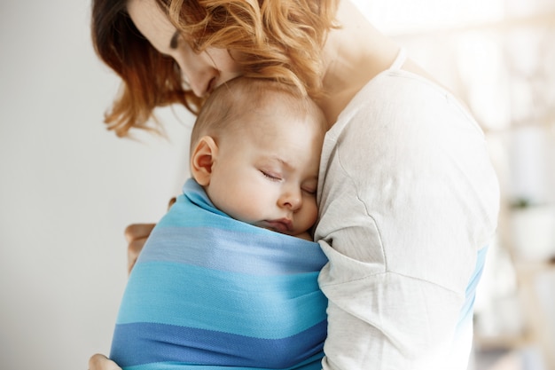 Kostbaar weinig pasgeboren jongen die diepe slaap hebben bij dag op moederborst in blauwe babyslinger. mamma die babyhoofd kust en ontspannen en verrukking voelt. familie concept.