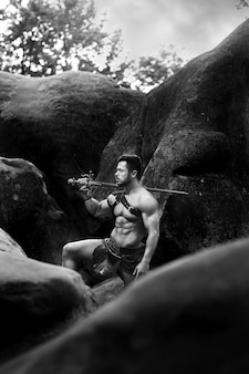 Krachtige mens. zwart-wit verticaal schot van een spartaanse krijger die bij de rotsen in het bos rust met een zwaard op zijn schouder