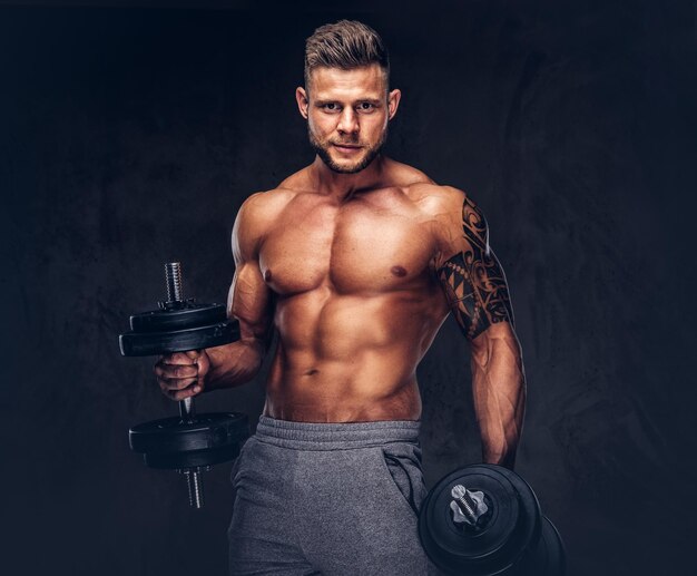 Krachtige stijlvolle bodybuilder met tatoeage op zijn arm, poseren met halters in een studio. Geïsoleerd op een donkere achtergrond.