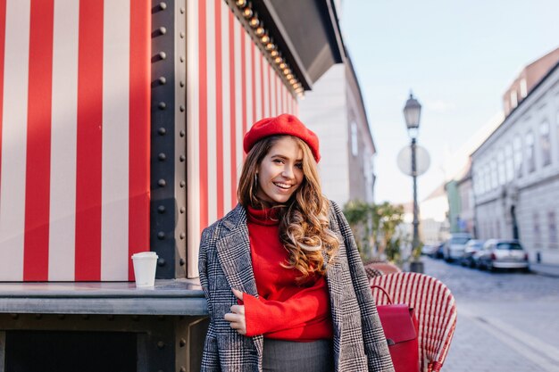 Krullend bruinharige meisje met oprechte glimlach poseren in grijze jas op mooie Europese straat