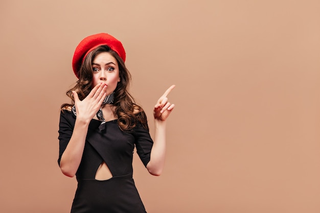 Krullend brunette meisje in zwarte jurk en rode baret bedekt haar mond van verbazing en wijst haar vinger naar rechts.