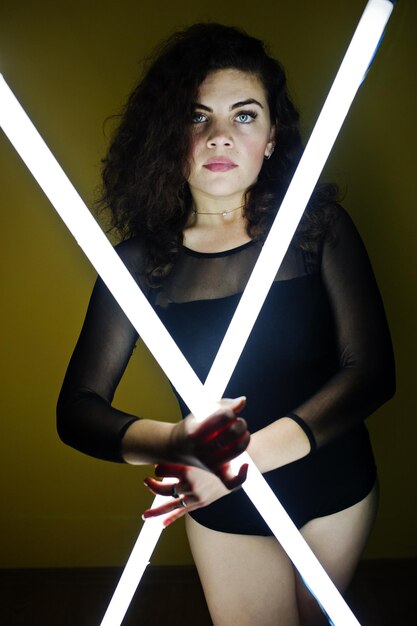 Krullend brunette plus size model met twee lange led-lampen tegen gele muur