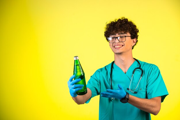 Krullend haarjongen in medisch uniform en handmaskers die een fles vloeistof tonen en glimlachen.