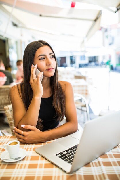 Krullend Latijns-meisje praten over de telefoon zittend op het terras. Vrij vrouwelijke freelancer in glazen koelen in restaurant.