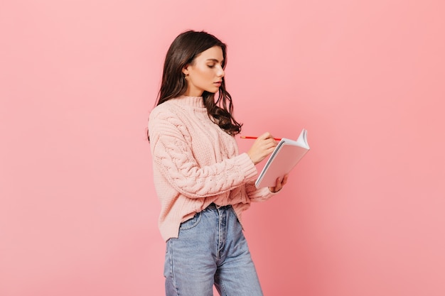 Krullend meisje maakt aandachtig aantekeningen in dagboek. vrouw in trui die zich voordeed op roze achtergrond.